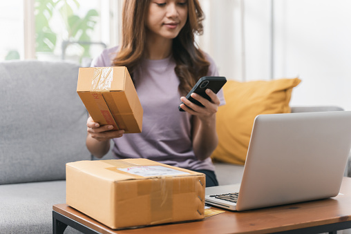 Portrait of cheerful young Asian woman using mobile phone for checking order and preparing products for delivery to online customer. Young woman receiving parcels with home delivery service