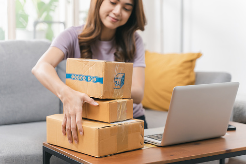 Portrait of cheerful young Asian woman using laptop for checking order and preparing products for delivery to online customer. Young business woman working e-commerce from home.