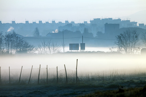 Silhouette of buildings emerging from a polluted environment with fog and smoke, close to a park with fences, electricity posts and signs.
