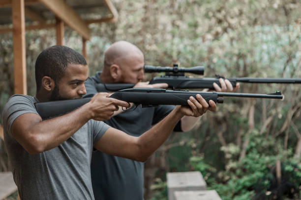 рыжий мужчина средних лет пытается стрелять из винтовки с младшим чернокожим инструктором - rifle range стоковые фото и изображения