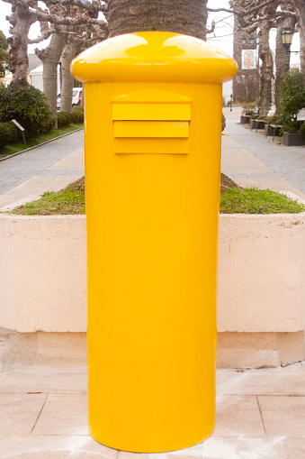 Close-up vertical view of yellow retro style mail box. Diminishing perspective of public park in the background. Navia, Asturias, Spain.