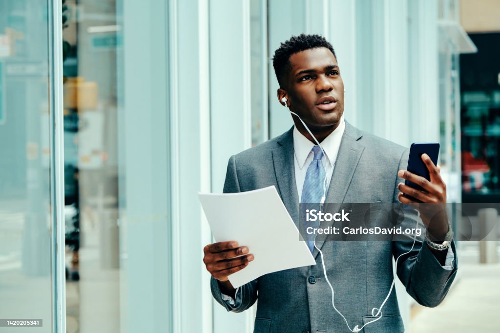 Young entrepreneur using smartphone outside wearing suit Young adult entrepreneur using smartphone outside wearing suit, tie and headphones holding documents looking away 20-24 Years Stock Photo