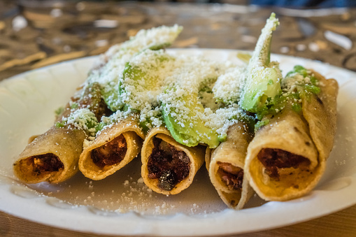 Close-up of mexican taquitos or flautas on plate with shredded cheese and avocado slices.