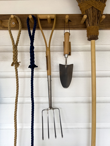 Vertical closeup photo of a collection of gardening tools hanging on a white painted timber wall.