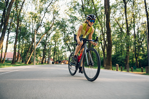 Young woman cyclist riding road bicycle on a free road in the forest at a sunny day. Healthy lifestyle concept. Wearing a protective helmet and an active wear while training for a race. Copy space.
