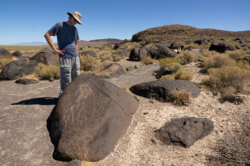 Pecked into volcanic basalt boulders at Grimes Point Archaeological Area in Nevada, a hiker explores the Great Basin rock art with circular and wavy lines dating from approximately 3000 years ago.