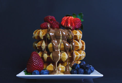 A stack of four waffles, with raspberries, blueberries and strawberries. Melted milk and white chocolate oozing down the side of the waffle stack.