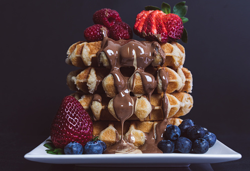 A stack of four waffles, with raspberries, blueberries and strawberries. Melted milk and white chocolate oozing down the side of the waffle stack.