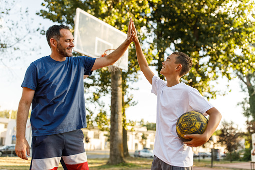 Candid shot of cheerful teenage boy high-fiving his father after they finished playing a great game of basketball together on the court at the sunny park.