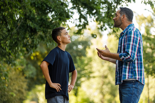 Side view of mid adult man gesturing while engaging in discussion and gently disciplining his teenage son, who is standing with hands on hips. They are standing on the city street, at the public park.