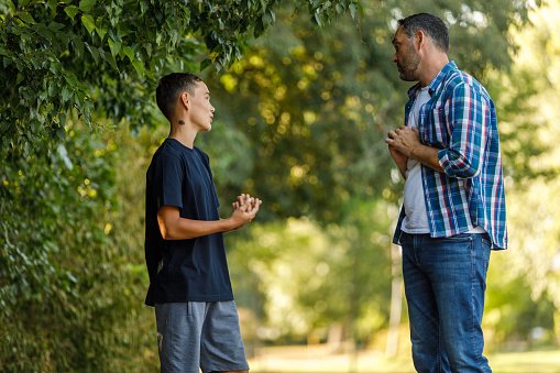 Side view of teenage boy and his father standing opposite each other at the public park, gesturing while engaging in an argument.