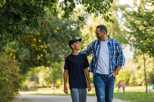 Copy space shot of contented mid adult man embracing his teenage son while they are taking a relaxing walk at the park. They are looking and smiling at each other, having a bonding moment.