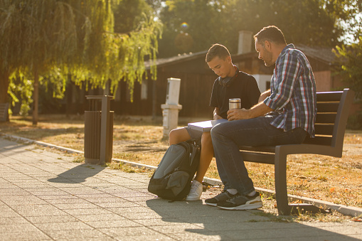 Wide shot of teenage boy opening a file binder with his schoolwork that he is showing to his dad while sitting on a park bench after school.