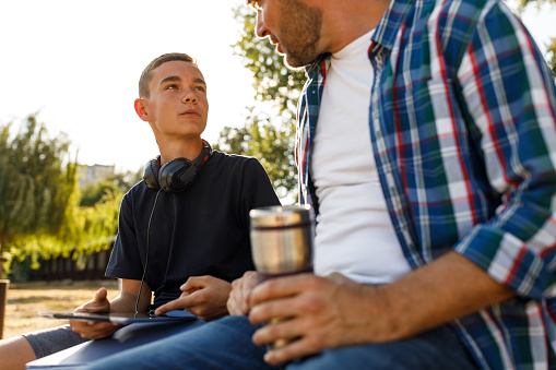Selective focus shot of mid adult man enjoying a cup of coffee and giving his teenage son some advice while talking about school and education while relaxing on a park bench.