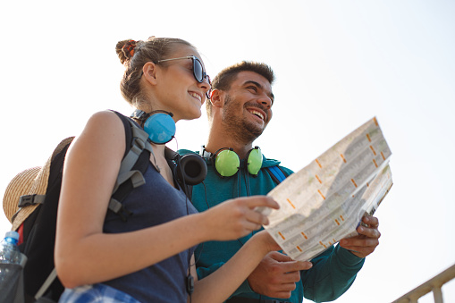 Low angle view of joyful heterosexual young couple holding a paper map, looking at the view and smiling.