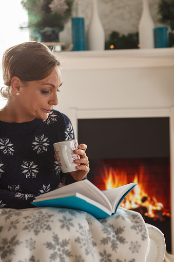 Portrait of happy mature woman sitting in armchair, by the fireplace, under a warm blanket, enjoying a cup of tea and reading a book.