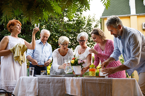 Candid shot of joyful group of senior friends standing around the table and setting up plates, cutlery, glasses and drinks for a garden dinner party.