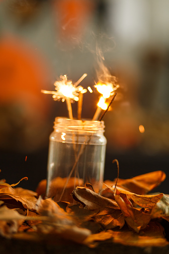 Close up shot of lit sparklers put in a glass jar, dried autumnal leaves scattered around for decoration.