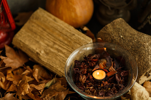 Copy space shot of a small tea candle burning in a bowl with potpourri, put in a pile of dried leaves, next to wooden logs as autumnal decoration.