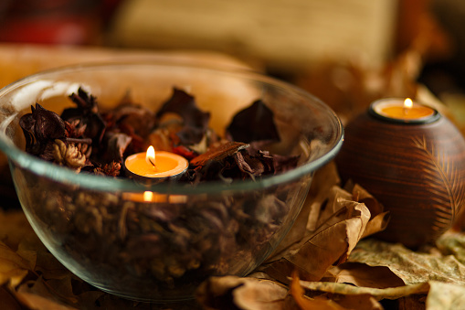 Close up shot of a glass bowl filled with scented potpourri and a tea candle burning in the middle, all arranged in a pile of dried leaves as autumnal decoration.