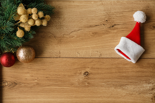 Directly above Christmas tree branch with sparkly ornaments and a small Santa's hat against a wooden background with copy space in the middle.