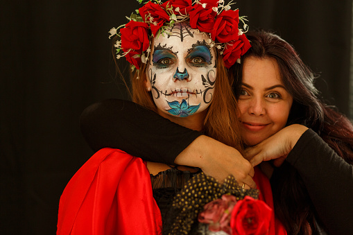 Portrait of joyful make up artist embracing her model in sugar skull face paint, from behind, both looking at camera and smiling.