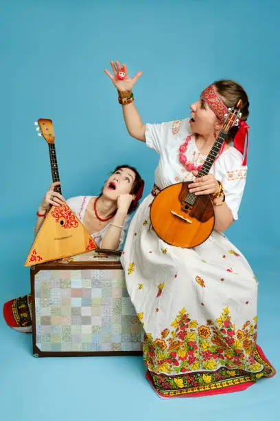 Photo of Women musicians in Russian folk dresses with musical instruments on a blue studio background. Happy artists from Russia in white national clothes with stringed musical instruments