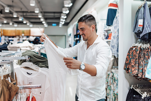 A young Caucasian man is holding a white t-shirt with a smile on his face, while taking a look around a clothing store.