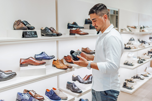 A young Caucasian man is holding a leather shoe in his hands while shopping in a modern shoe store.