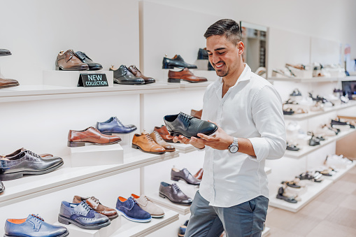 A young Caucasian man is standing in a modern shoe store and taking a look at a leather shoe with a smile on his face.