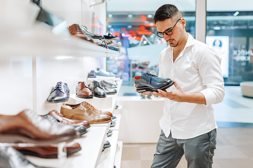A young Caucasian man wearing eyeglasses is holding a leather shoe in his hand and taking a look at it while shopping in a store.