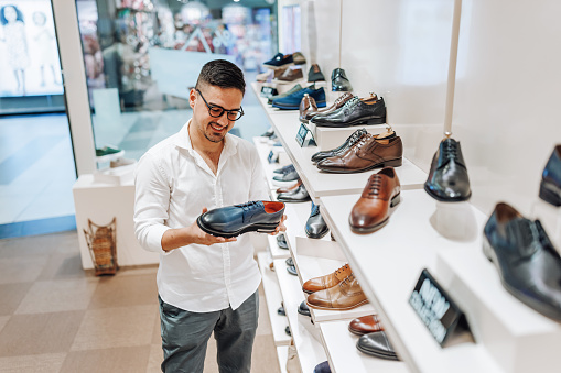 A young Caucasian man is standing in a modern shoe store, taking a look at a pair with a cheerful expression on his face.