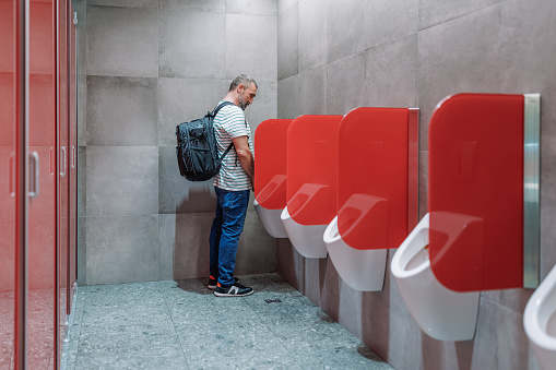 A mature Caucasian male tourist wearing a black backpack is using the public restroom.
