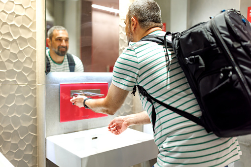 A mature Caucasian male tourist is looking at himself in the mirror with a smile, while washing his hands in a public restroom.