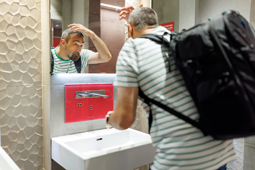 A mature Caucasian male tourist is touching up his hairstyle in the public restroom mirror.