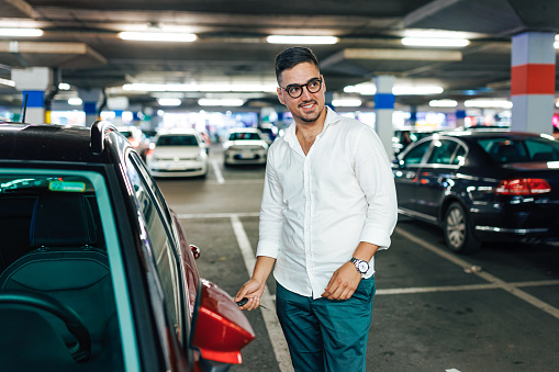 A young Caucasian man is looking away with a smile on his face while standing next to his car in an underground garage.