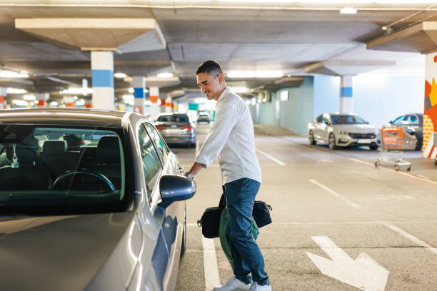 Can't wait to start my trip A young Caucasian man is holding a bag in his hand and opening his car door with a smile on his face, while standing in an underground garage. parking stock pictures, royalty-free photos & images