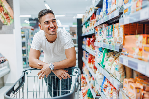 A young Caucasian man is leaning against a shopping cart and taking a look around the supermarket with a big smile on his face.