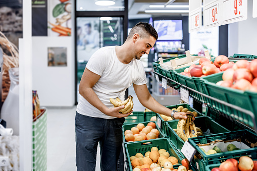 A young Caucasian man is leaning and picking up bananas with a smile on his face while shopping in a supermarket.
