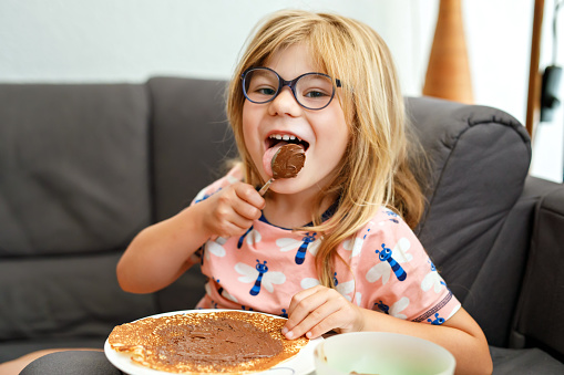 Adorable little girl having breakfast, eating pancakes with chocolate cream. Preschool child smiling. Sweet food for children