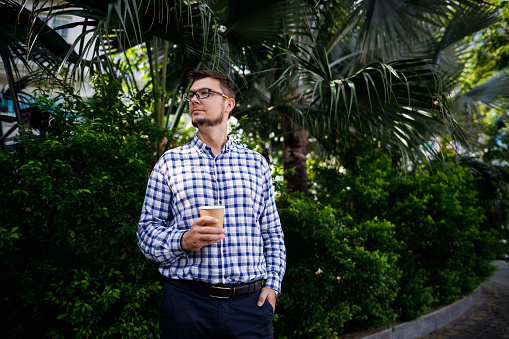 Handsome businessman holding a cup of coffee, wearing a plaid shirt, glasses, with a trimmed beard standing on the street looking away in Tay Ho, Hanoi Vietnam