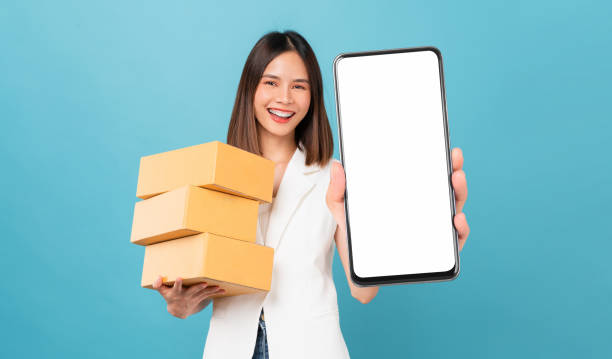 Smiling beautiful Asian woman holding cardboard boxes and hands show smartphone mockup of blank screen on blue background. Take your screen to put on advertising. Concept delivery online. stock photo