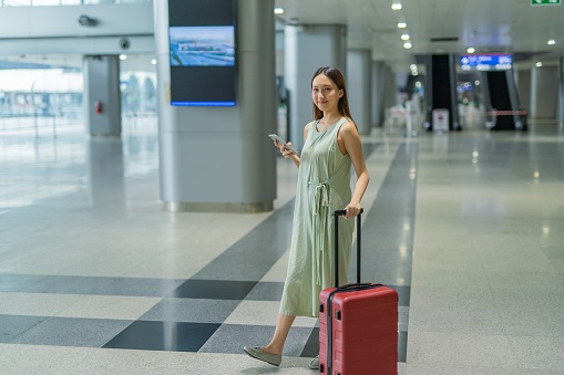 Portrait photo of a young asian female lady with her phone and suitcase preparing and going for a travel to her dream holiday destination. She is constantly checking her phone and chatting with her friends to share her happiness.