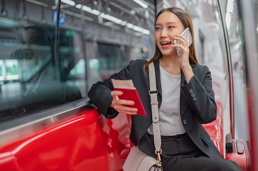 Portrait photo of a young asian businesswoman lady traveling in a commuter rail train with a red luggage going for a business trip, chatting to her colleague on her phone a