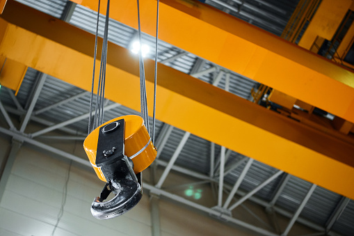 Huge hook assembly of bright yellow moving crane hangs under ceiling in workshop of production plant close low angle shot
