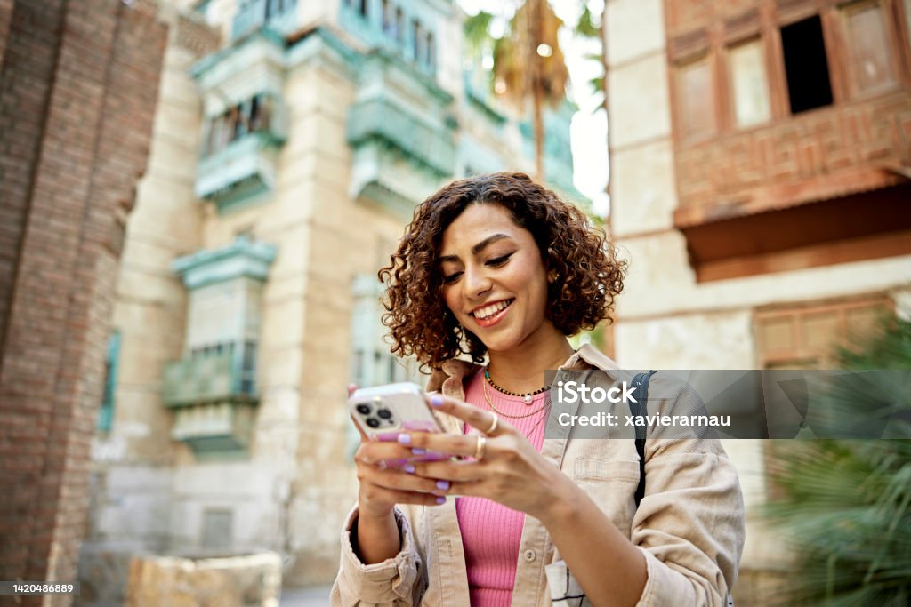 Candid portrait of young Middle Eastern digital native Waist-up view of casually dressed woman with curly brown hair looking at smart phone and smiling while visiting historic Al-Balad, Jeddah. People Stock Photo