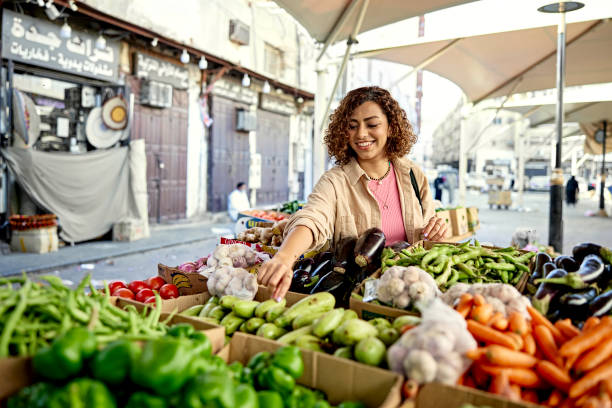 Young woman grocery shopping at outdoor market Waist-up view of smiling Middle Eastern woman in casual attire selecting zucchini from variety of vegetables in retail display under protective umbrellas. jeddah stock pictures, royalty-free photos & images