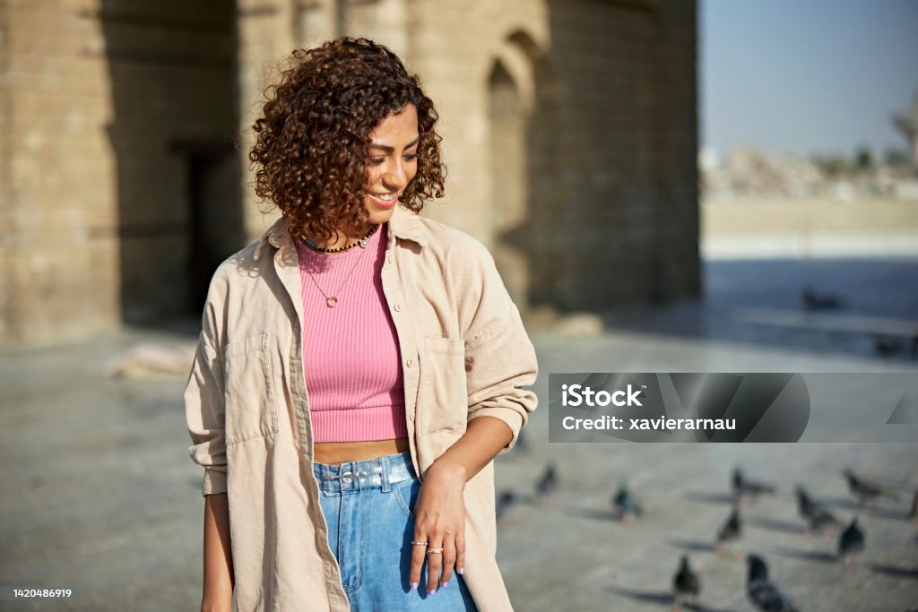 Relaxed young woman outdoors at Baab Makkah in Jeddah Front view of cheerful Middle Eastern woman in casual attire looking down as she stands in sunshine with historical landmark and pigeons in background. Adult Stock Photo
