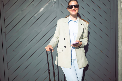 A portrait of a smiling woman looking directly at the camera while standing next to her suitcase.