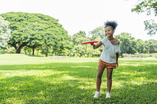 Happy African American little girl with curly hair has fun playing with red flying disc in the park. kid activity outdoor.